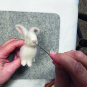 Ein quadratisches Stück Filz liegt auf einem weißen Tisch. Darauf sieht man zwei Hände, die gerade die Nase des kleinen Kaninchens bearbeite.