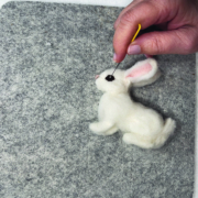 Ein quadratisches Stück Filz liegt auf einem weißen Tisch. Darauf sieht man eine Hand, die gerade das Auge des Kaninchens anfilzt