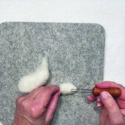 Ein quadratisches Stück Filz liegt auf einem weißen Tisch. Darauf sieht man zwei Hände, die ein kleiners Stück Wolle mit einer Filznadel bearbeiten. Danben liegt ein größeres Stück Wolle.