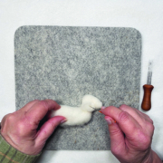 Ein quadratisches Stück Filz liegt auf einem weißen Tisch. Darauf sieht man zwei Hände, die gerade den Kopf des Kaninchens mit einer Filznadel bearbeiten.