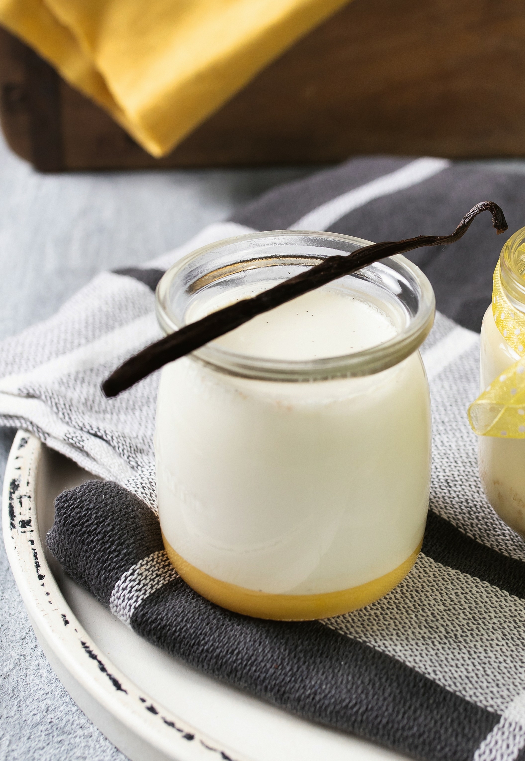 Ein durchsichtiges kleines Gläschen, in dem sich ganz unten eine Schicht Lemon Curd befindet und darüber das Vanillejoghurt. Eine Vanilleschote liegt als Dekoration oben auf dem Gläschen.
