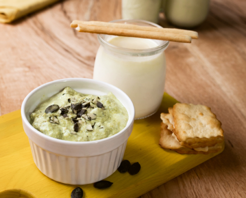 Auf einem gelben Holzbrett steht ein kleines weißes Schälchen mit Bärlauch-Kürbiskern-Frischkäse. Daneben steht ein kleines Glas mit Joghurt. Ein paar Cracker sowie Kürbiskerne liegen als Deko daneben.