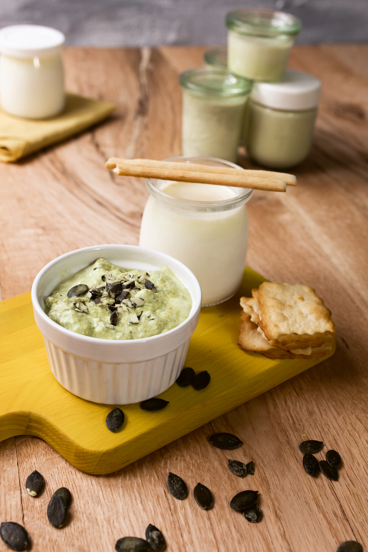 Auf einem gelben Holzbrett steht ein kleines weißes Schälchen mit Bärlauch-Kürbiskern-Frischkäse. Daneben steht ein kleines Glas mit Joghurt. Ein paar Cracker sowie Kürbiskerne liegen als Deko daneben.