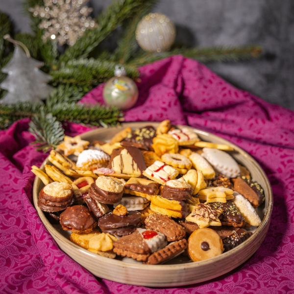 1 Ein Teller mit vielen verschiedenen Kekssorten. Dahinter Tannenzweige und Weihnachtskugeln und -dekoration