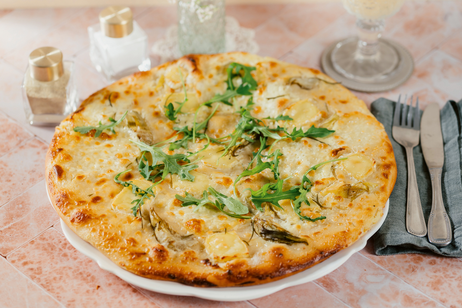23Hö_Pizza bianca ganz Die Pizza liegt auf einem Teller, garniert mit Ruccola. Daneben liegen Messer und Gabel auf einer Serviette.