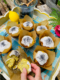 Mehrere Zitronen-Mohn-Muffins, die auf einem bunten Tablett angerichtet wurden. Eine Zitrone als Deko liegt daneben.