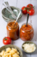 Zwei Glasgläschen gefüllt mit Sugo stehen nebeneinander auf einem Tisch. Daneben sind zwei Schälchen gefüllt mit Nudeln, und Parmesan, sowohl zwei leere Schälchen, in denen zwei Gabeln liegen.