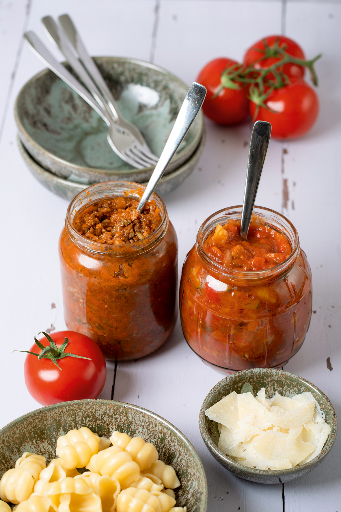 Zwei Glasgläschen gefüllt mit Sugo stehen nebeneinander auf einem Tisch. Daneben sind zwei Schälchen gefüllt mit Nudeln, und Parmesan, sowohl zwei leere Schälchen, in denen zwei Gabeln liegen.