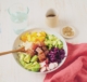 Ein Schüssel, in der Reis, Mango, Gurken, Rotkraut, Radieschen, Avocado und Lachs liegen. Daneben steht ein Wasserglas, ein kleines Schüsselchen mit Sesam.